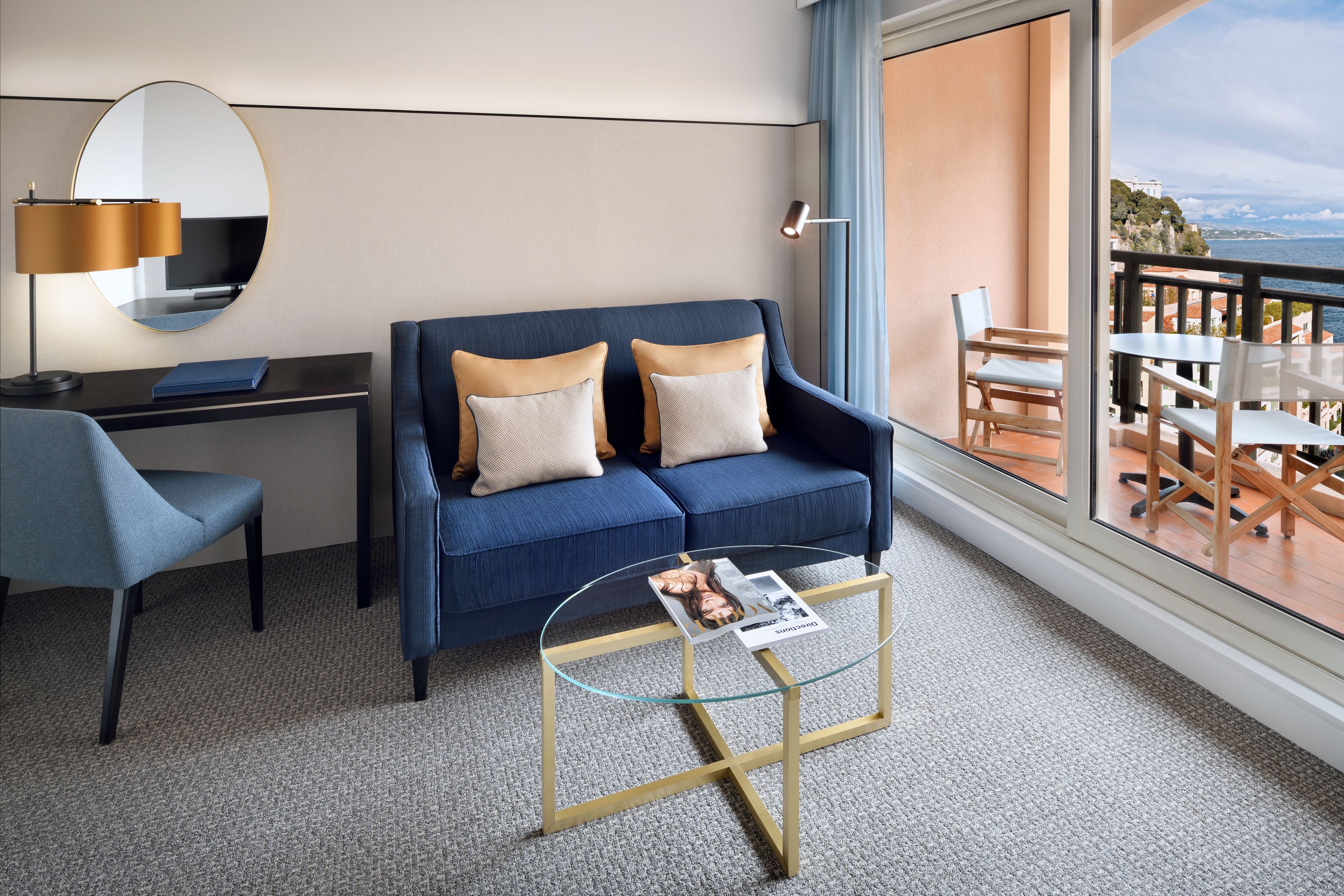 Ein modernes Hotelzimmer mit einem blauen Sofa, einem Schreibtisch, einem Glastisch und Meerblick.