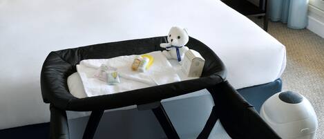 Une chambre d’hôtel avec un lit, une table de chevet avec une lampe, une petite table avec un vase de fleurs, et une poussette pour bébé avec un siège pour bébé et un ours en peluche.