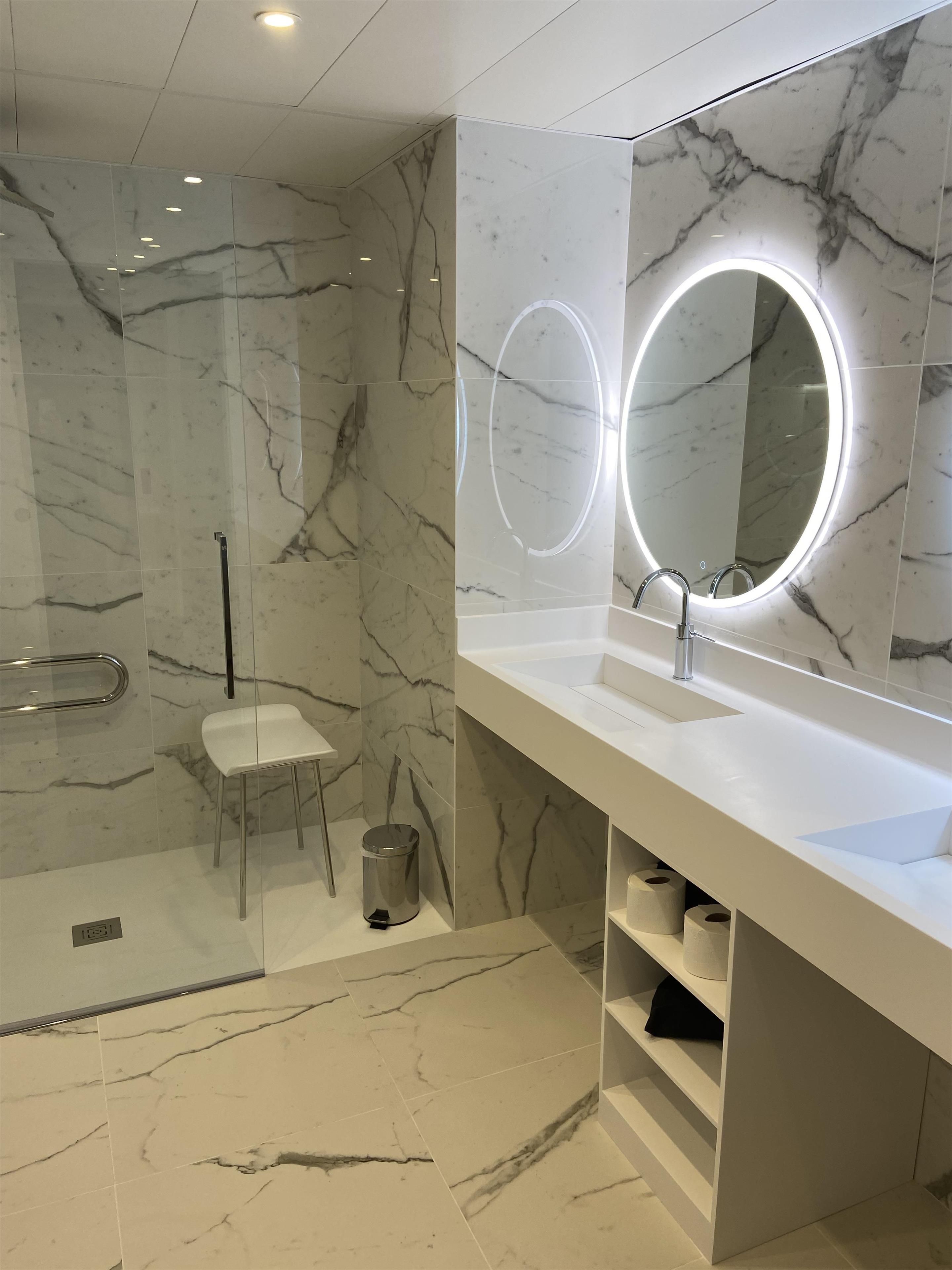 Une salle de bain moderne avec un sol en marbre, un lavabo blanc agrémenté d’un miroir rond et une cabine de douche en verre.