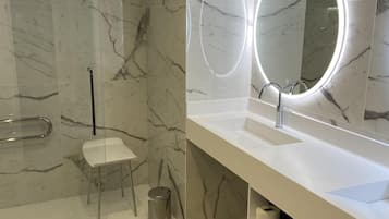 Une salle de bain moderne avec un sol en marbre, un lavabo blanc agrémenté d’un miroir rond et une cabine de douche en verre.