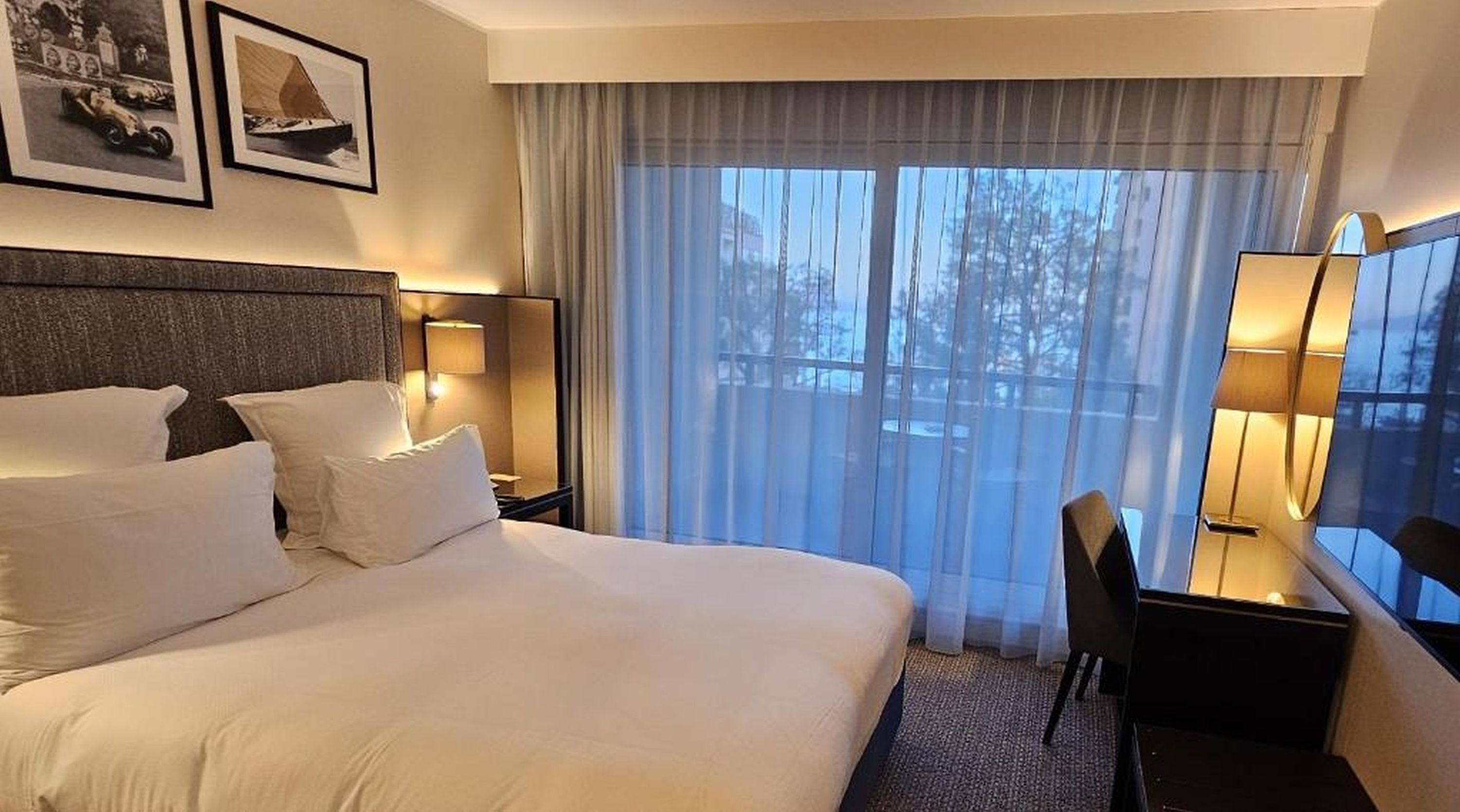 Een hotelkamer met een groot bed, twee bedlampen, een bureau met stoel, en een raam met gordijnen.