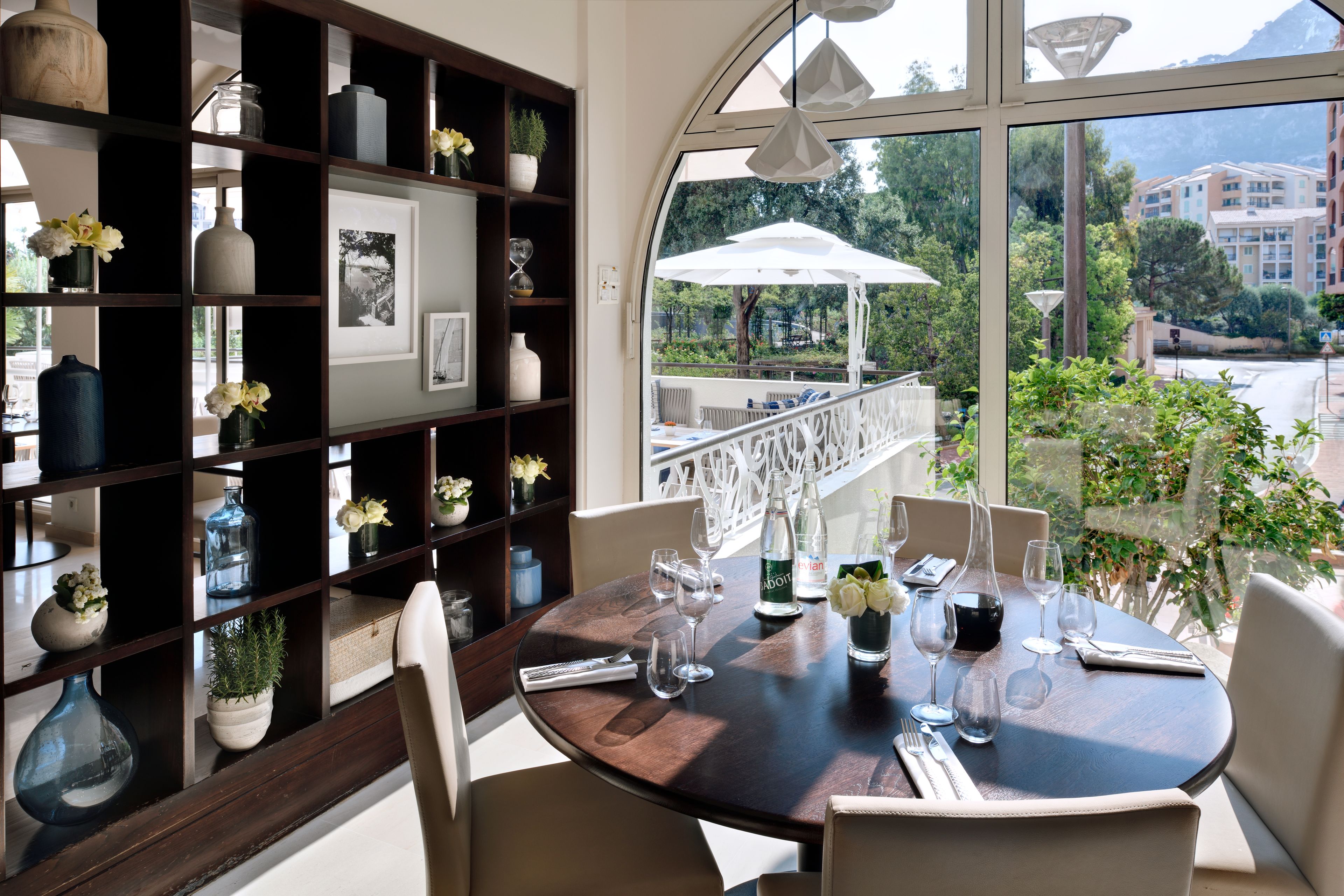 A round dining table with chairs, a bookshelf with decorative items, and a view of a garden and buildings through large windows.