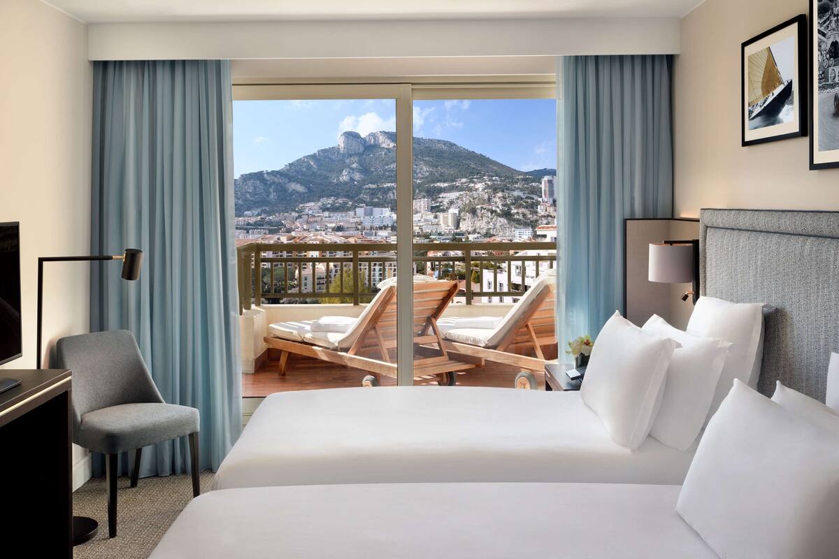 Ett hotellrum med en stor säng, ett skrivbord, en stol och en balkong med utsikt över ett berg och stadsbilden.