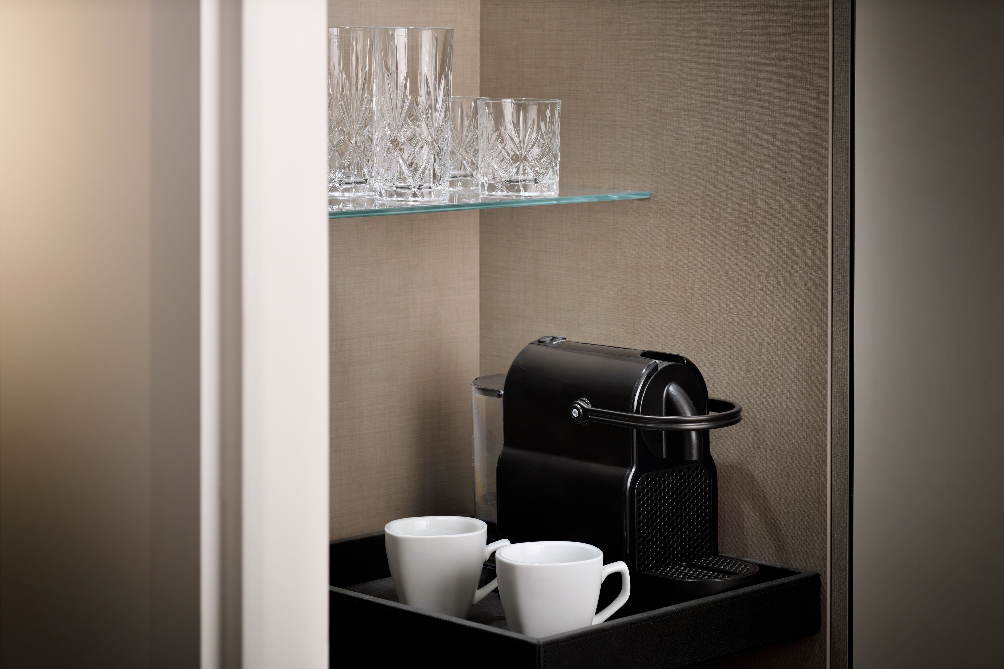 A coffee machine with a tray of two white cups, a glass shelf with multiple clear glasses, and a textured wall.