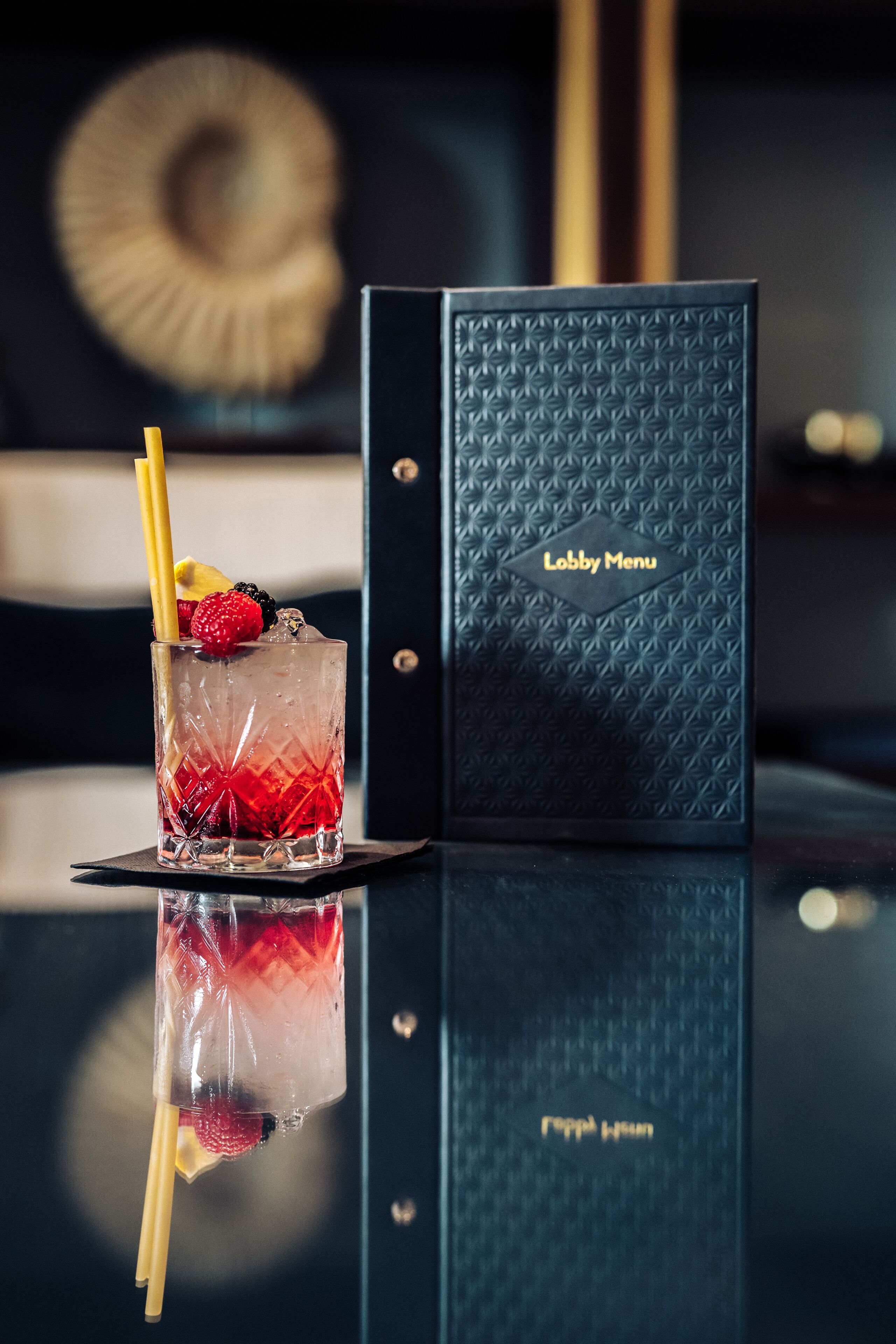A cocktail glass with fruit garnish on a reflective surface, a black menu with 'Lobby Menu' text, and a blurred background with a circular object.