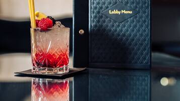 A cocktail glass with fruit garnish on a reflective surface, a black menu with 'Lobby Menu' text, and a blurred background with a circular object.