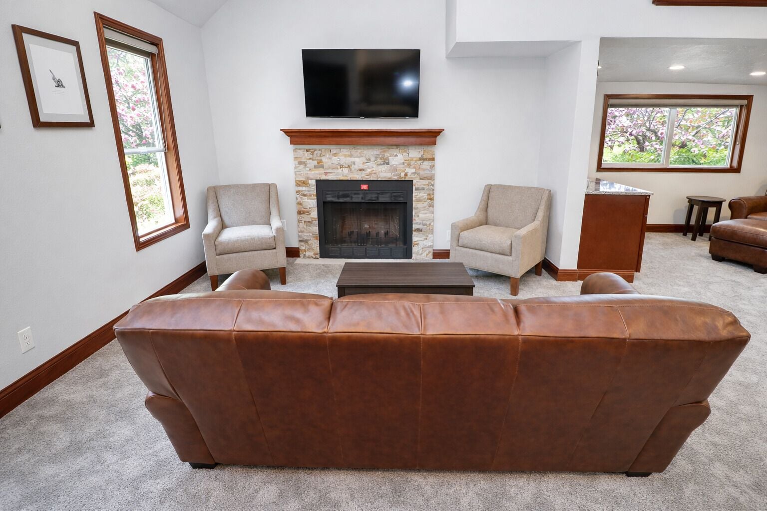 A living room with a stone fireplace, two armchairs, a leather sofa, a TV mounted above the fireplace, and a window with a view of trees.