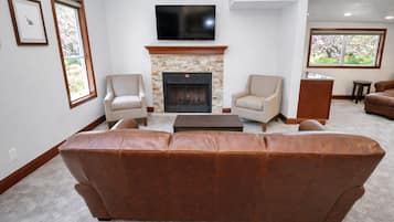 Un salon doté d’une cheminée en pierre, de deux fauteuils, d’un canapé en cuir, d’un téléviseur fixé au-dessus de la cheminée et d’une fenêtre donnant sur la forêt.
