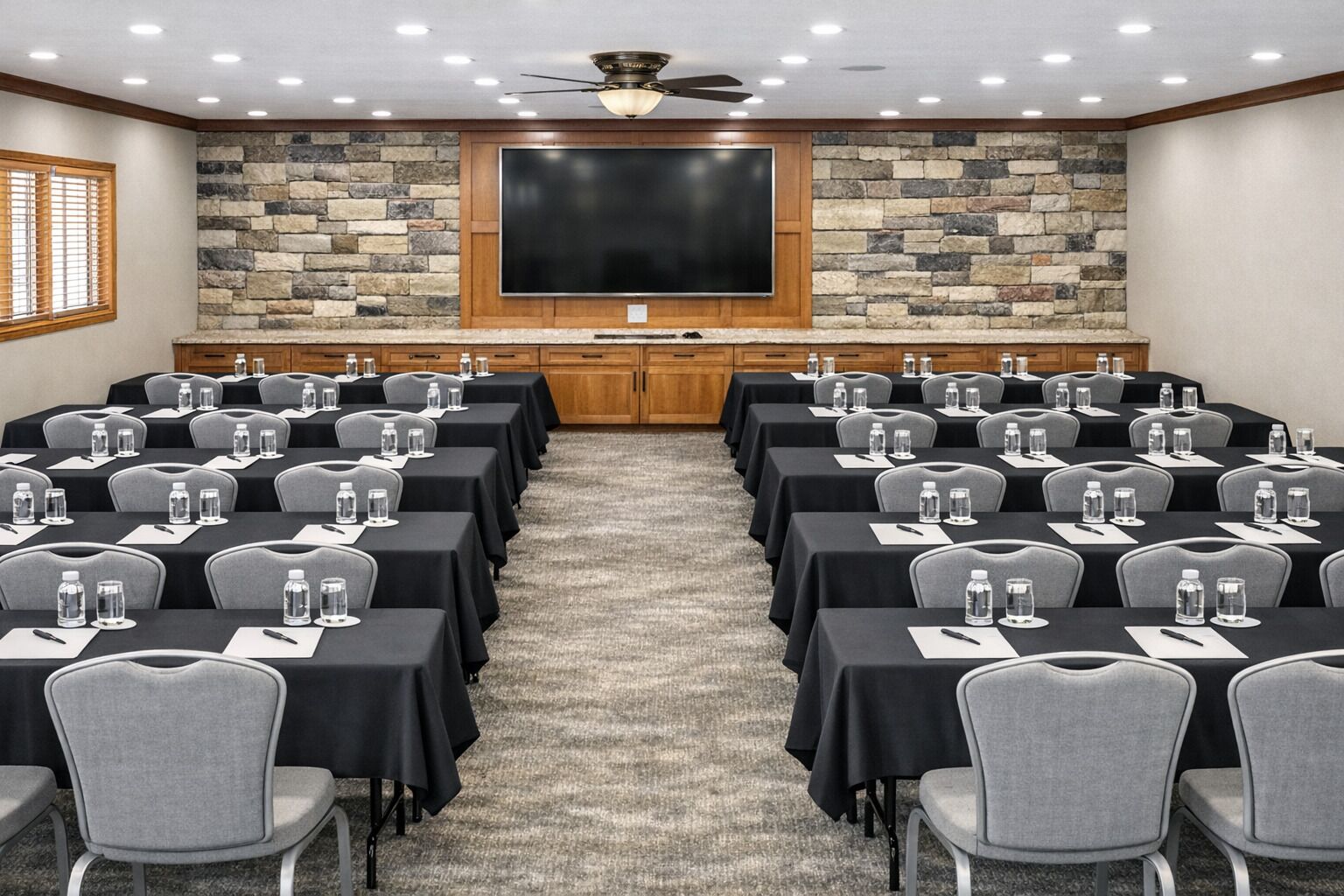 Un salón de conferencias con varias mesas, sillas, un televisor de pantalla plana grande, una pared de acento de piedra y un ventilador de techo.
