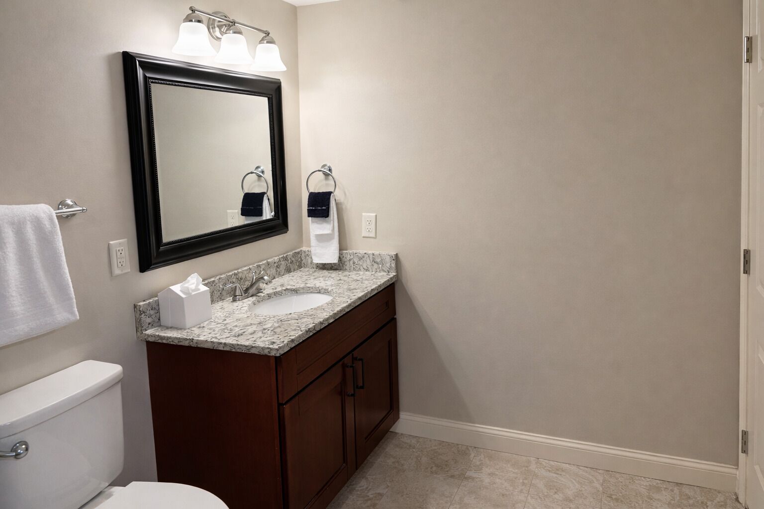 A bathroom with a white toilet, a marble sink, a dark wooden vanity, a large mirror with three lights, and a towel rack.