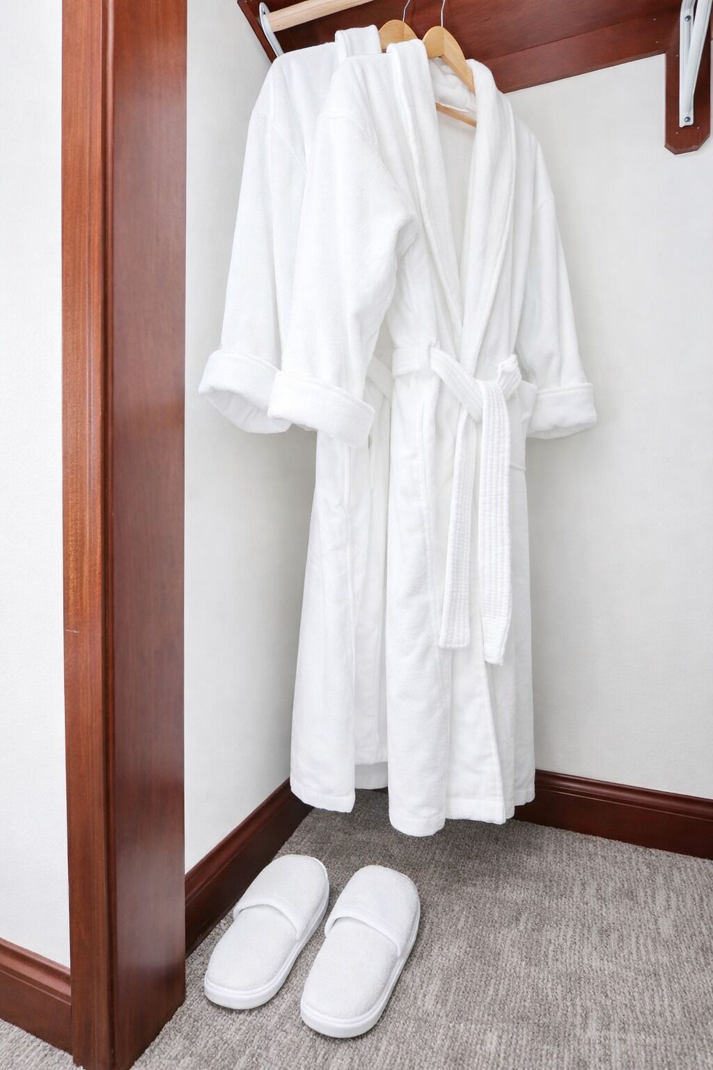 A white bathrobe hanging on a wooden hanger in a corner with a pair of white slippers on the floor.