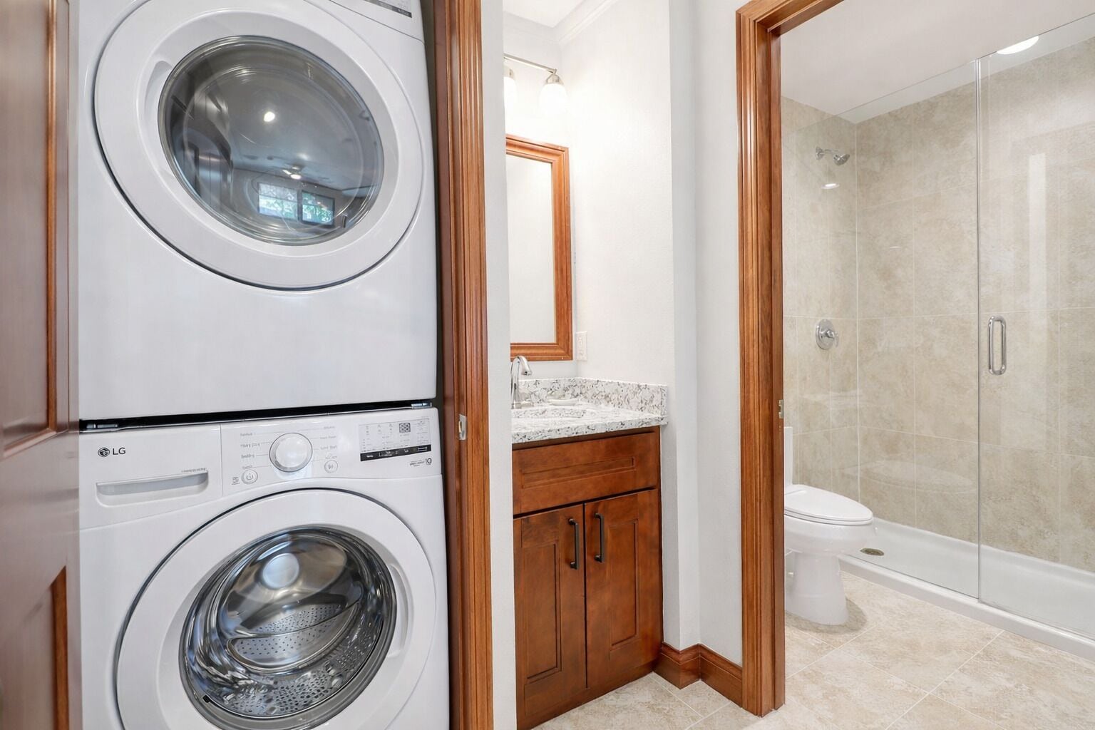A laundry area with two stacked washing machines, a wooden cabinet, a marble countertop, and a glass-enclosed shower.