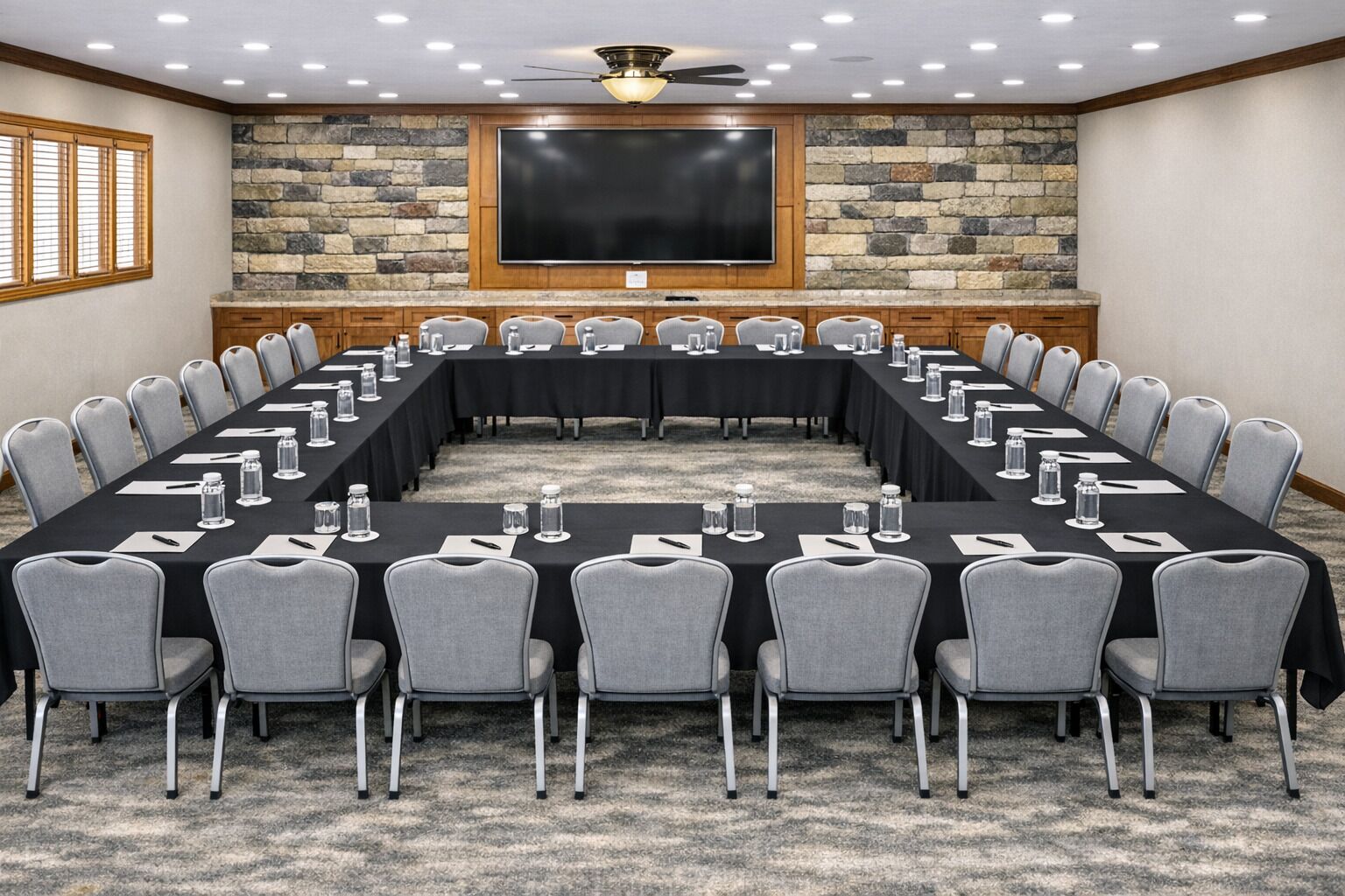 A conference room with a long table, grey chairs, a large screen, and a stone wall.