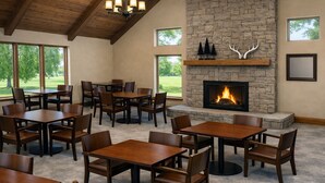 A spacious indoor area with multiple wooden tables and chairs, a stone fireplace, and large windows offering a view of greenery.