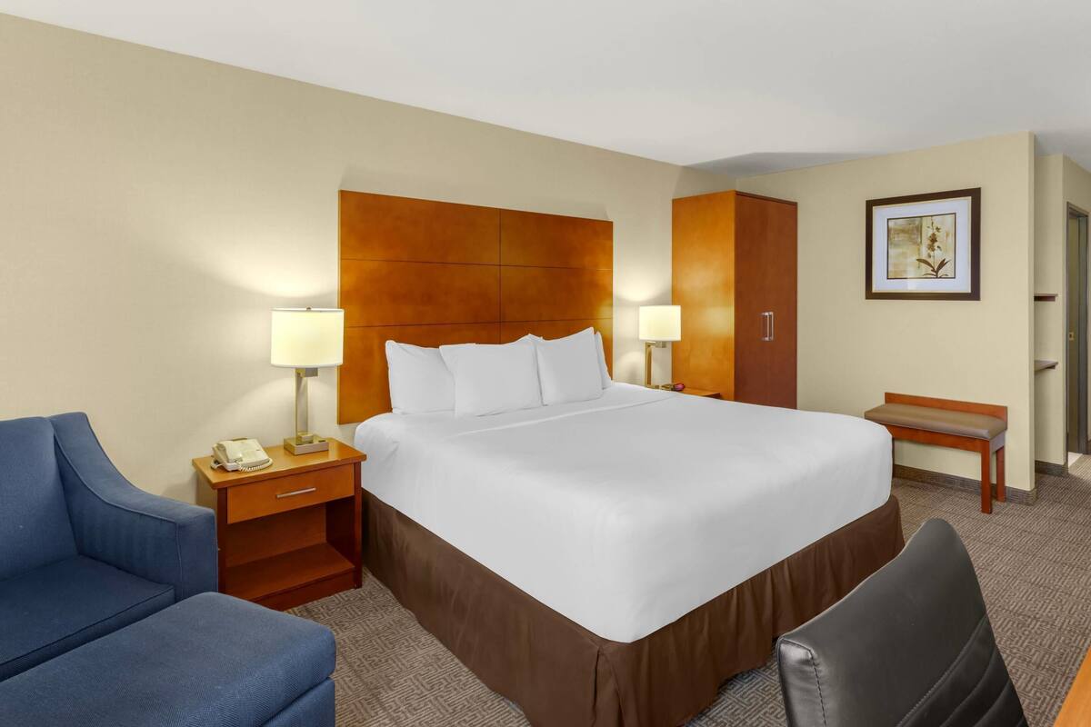 A hotel room with a large bed, a desk with a lamp, a blue armchair, and a wooden bench.