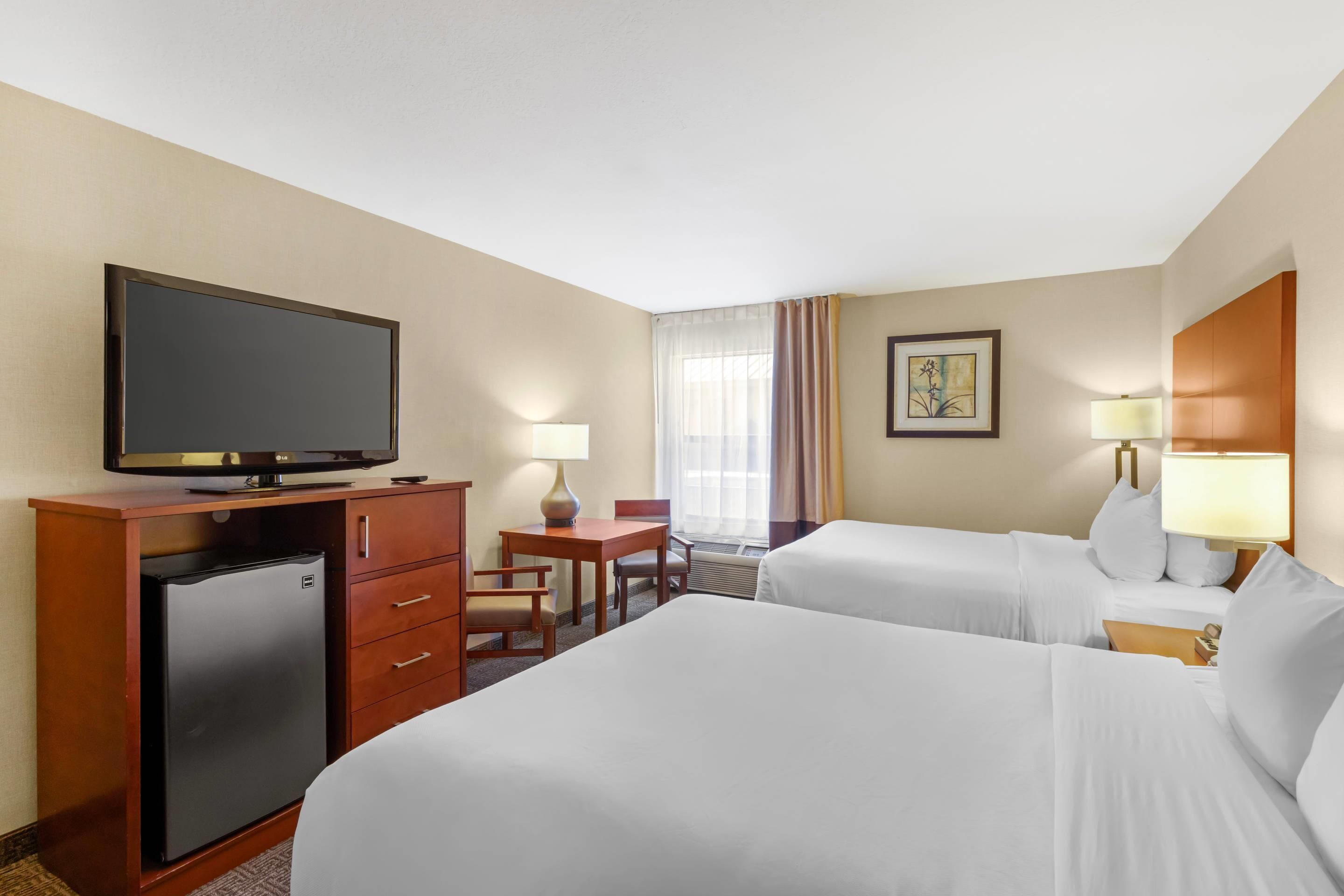 A hotel room with two beds, a television, a desk with a lamp, and a refrigerator.