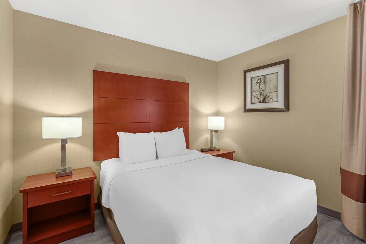 A hotel room with a large bed, two bedside tables with lamps, a wooden headboard, a framed picture on the wall, and a curtain on the window.