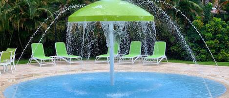 A pool with a central water fountain and four green lounge chairs around it.