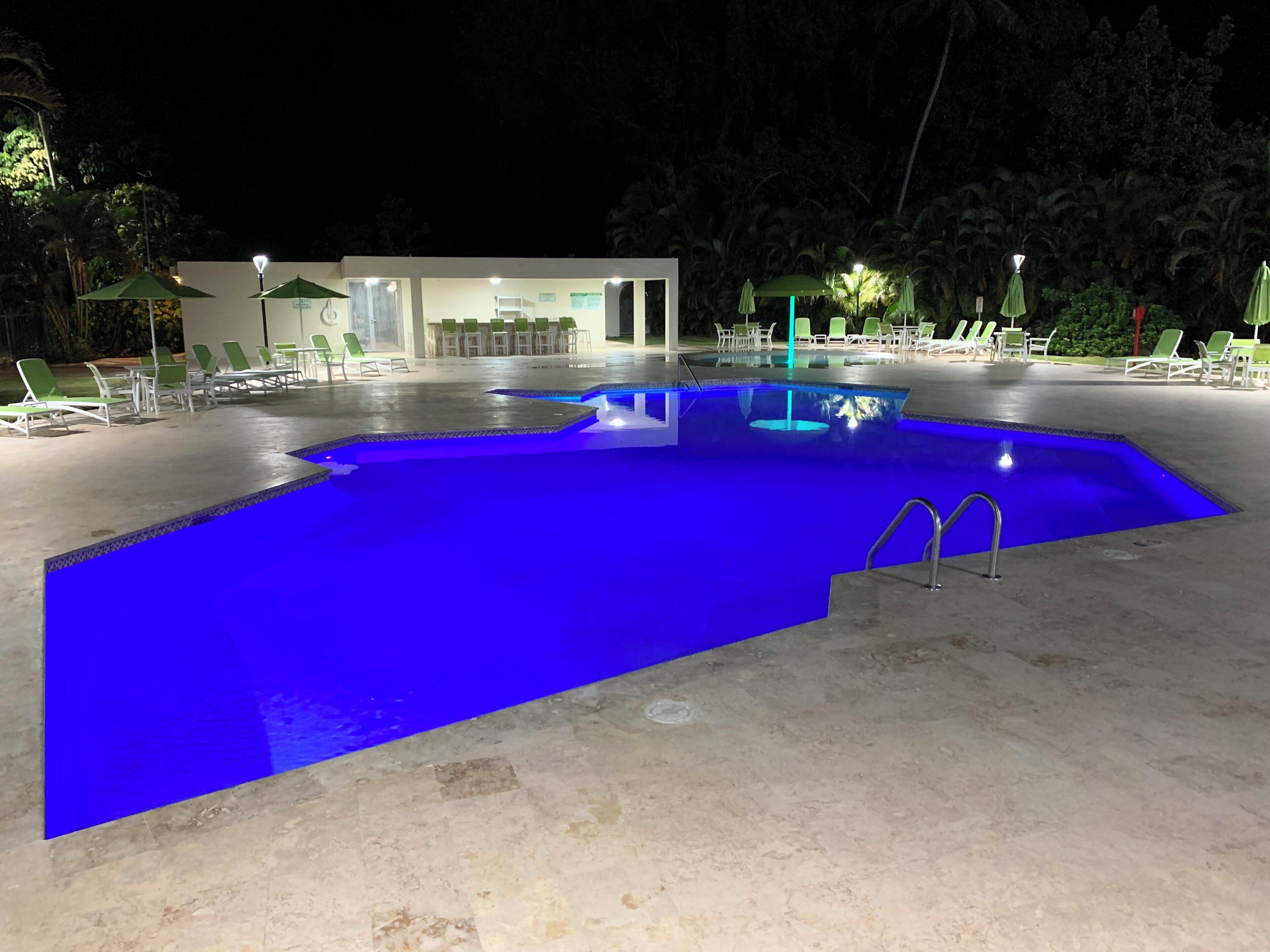 Ein Swimmingpool mit blauer Färbung, umgeben von Liegestühlen und Sonnenschirmen, mit einem Gebäude im Hintergrund.