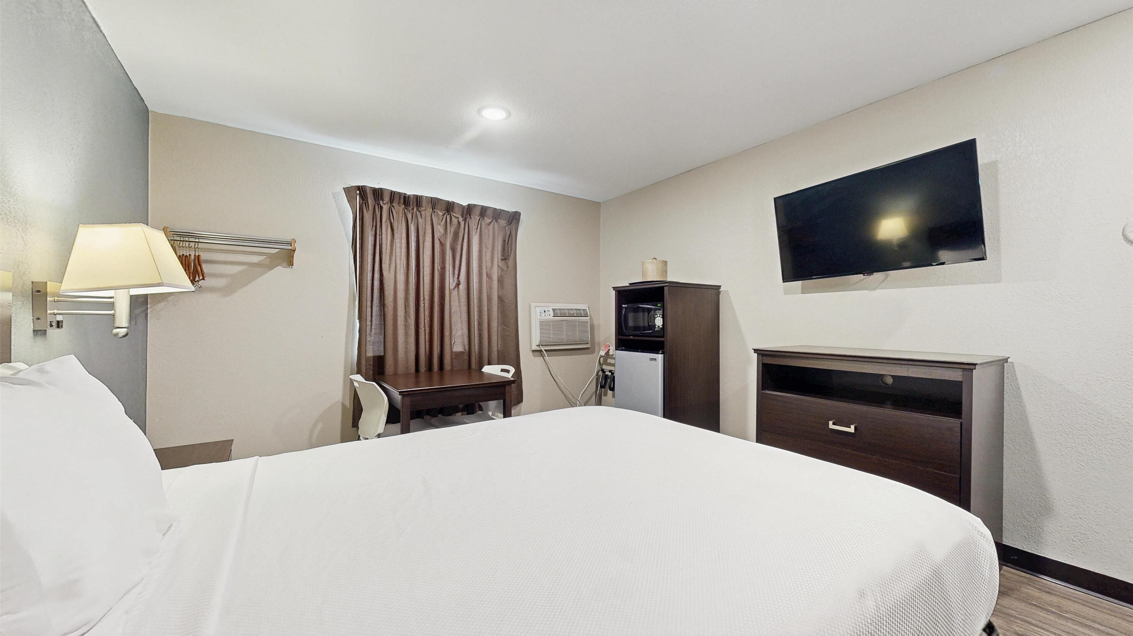 A hotel room with a bed, a desk, a chair, a TV, a microwave, and an air conditioning unit.