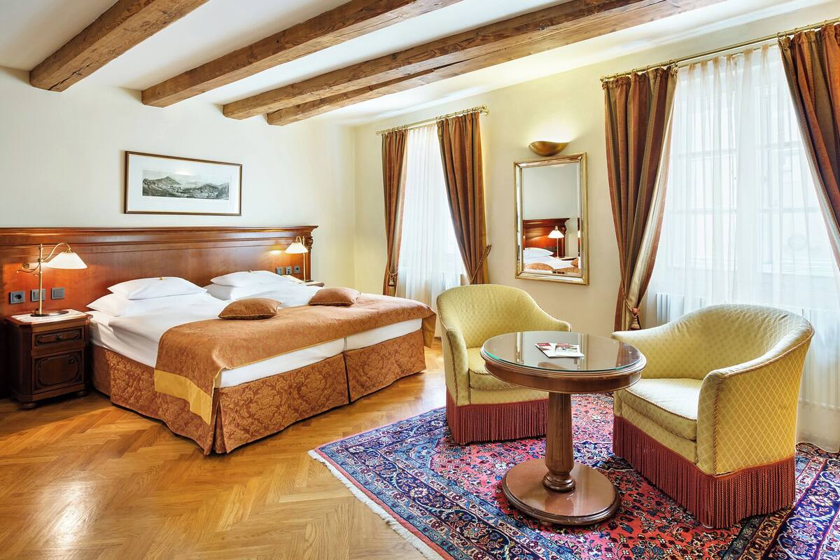A hotel room with a large bed, wooden beams on the ceiling, a round table, two armchairs, a patterned rug, and a large mirror.