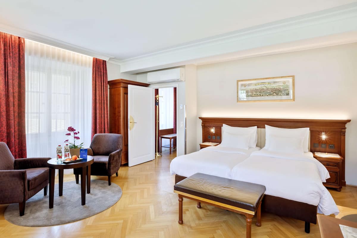 A hotel room with a large bed, two bedside tables, a wooden bench, a round table with chairs, and a large window with curtains.
