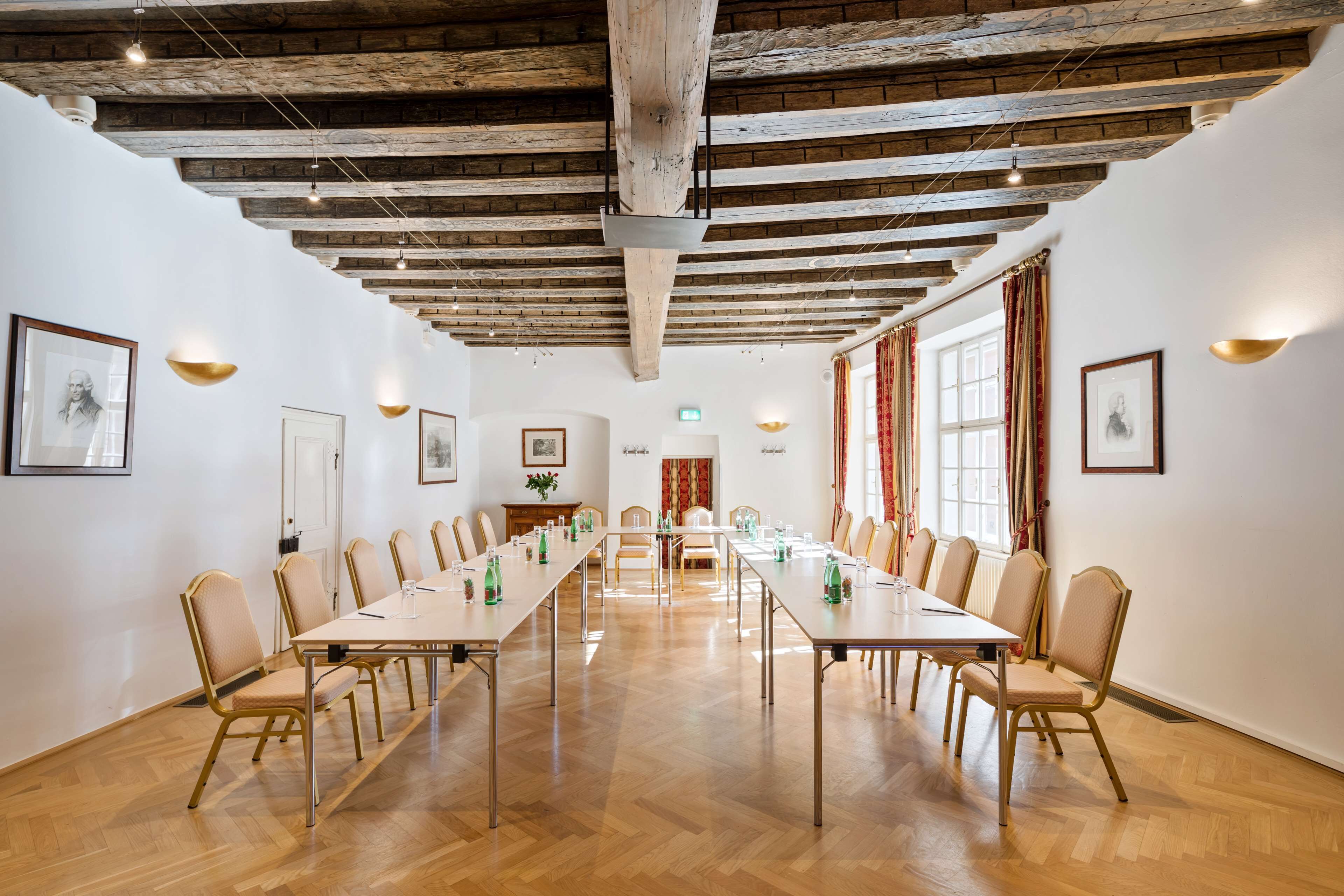 Un salón de conferencias con mesas largas y sillas, vigas de madera en el techo y cuadros en las paredes.