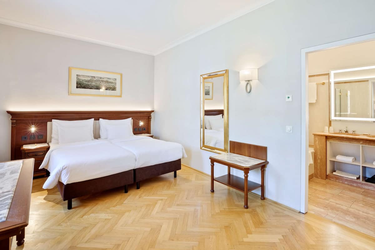 A hotel room with a double bed, a wooden nightstand, a bedside table, a chair, a large mirror, and a bathroom with a vanity and shelves.