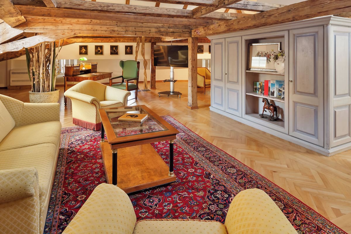 A spacious living room with a high wooden ceiling, a large wooden cabinet, a green armchair, a beige sofa, and a red patterned rug.