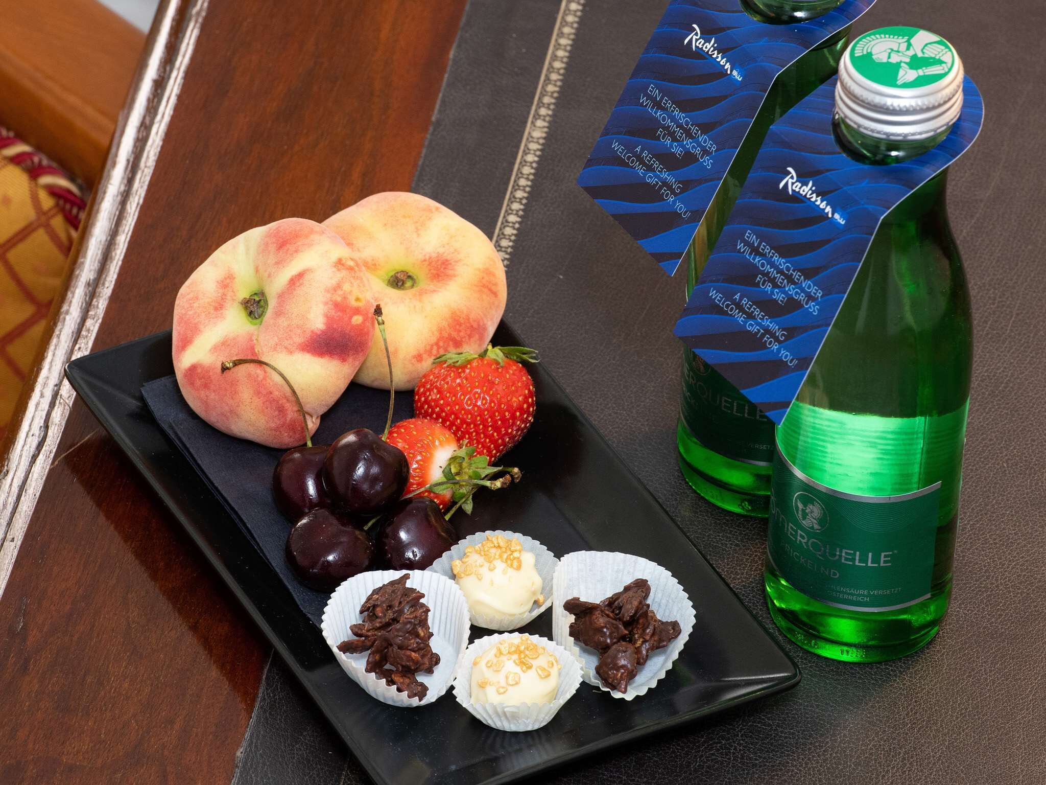 Un plato con melocotones, fresas, cerezas y nueces cubiertas de chocolate, junto a una botella de bebida verde y una caja con etiqueta azul.
