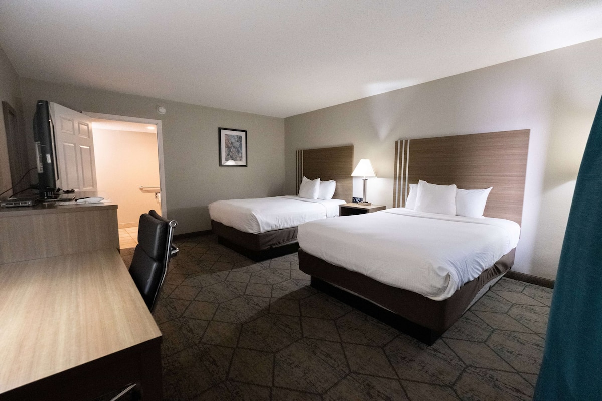 A hotel room with two beds, a desk, a chair, a TV, and a lamp.