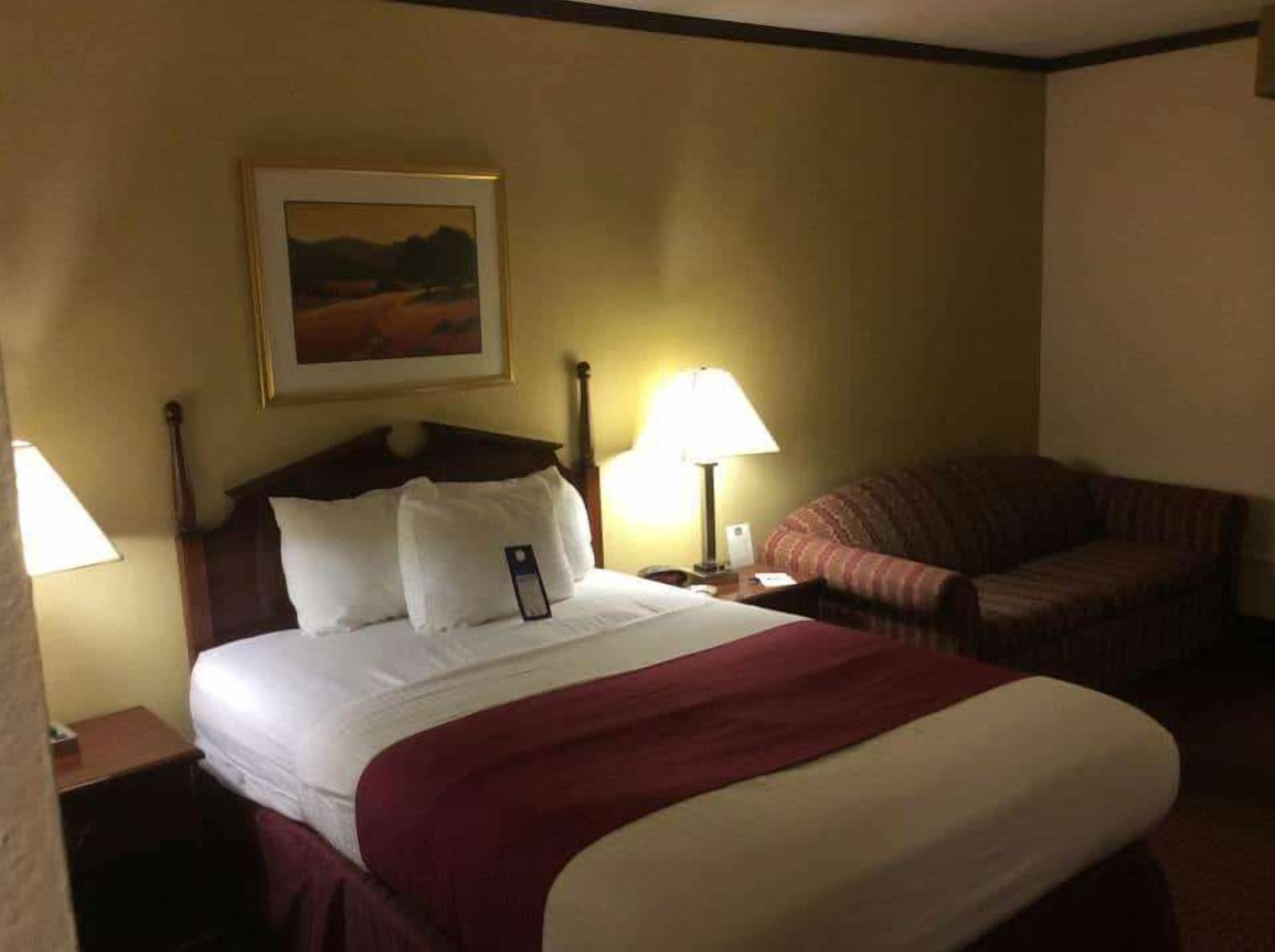 A hotel room with a bed, a nightstand, a lamp, a chair, and a painting on the wall.