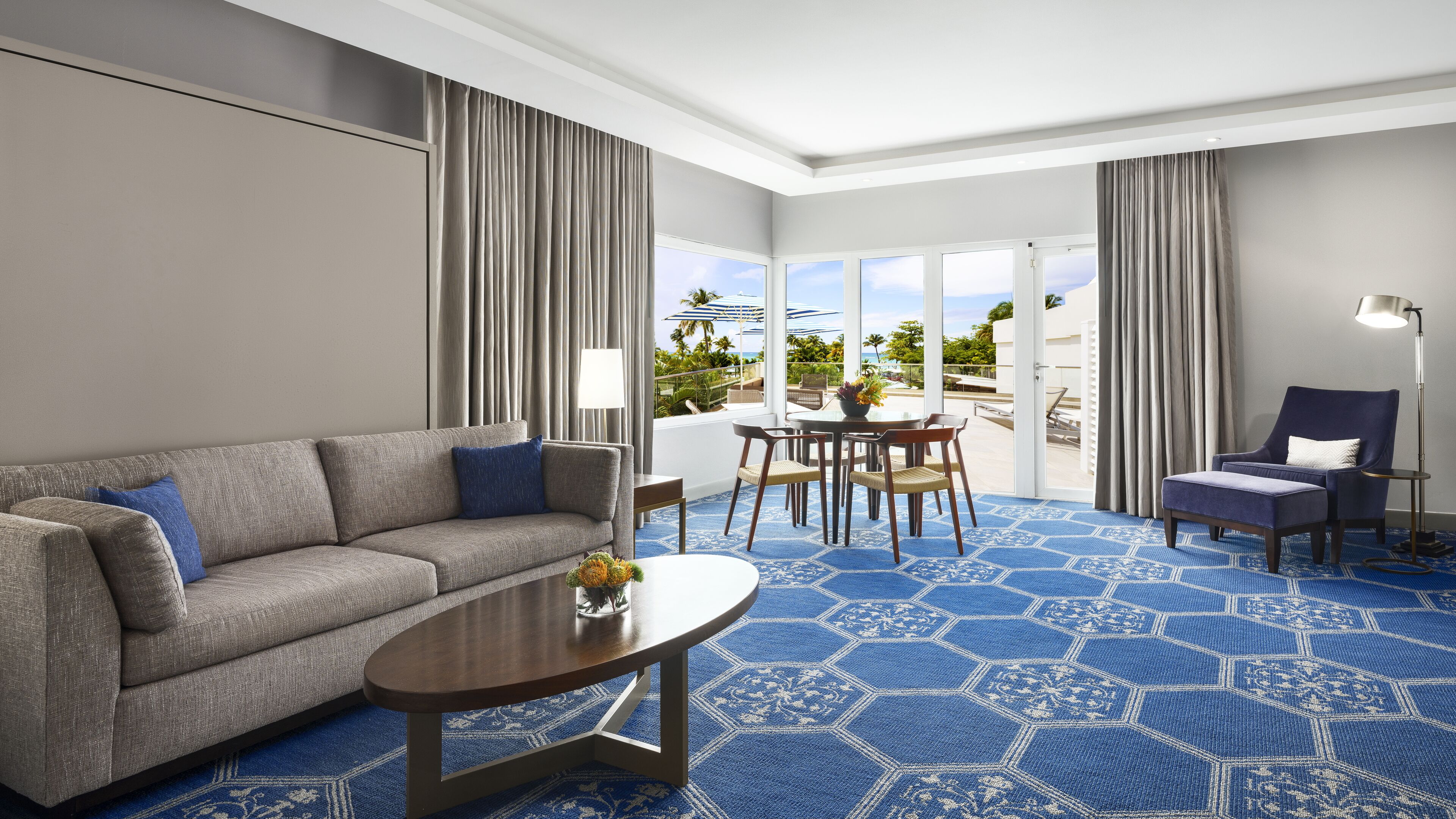 Habitación de hotel con sofá gris, una mesa redonda, zona de comedor con mesa y sillas, alfombra de estampado azul y vistas al exterior a través de amplios ventanales.