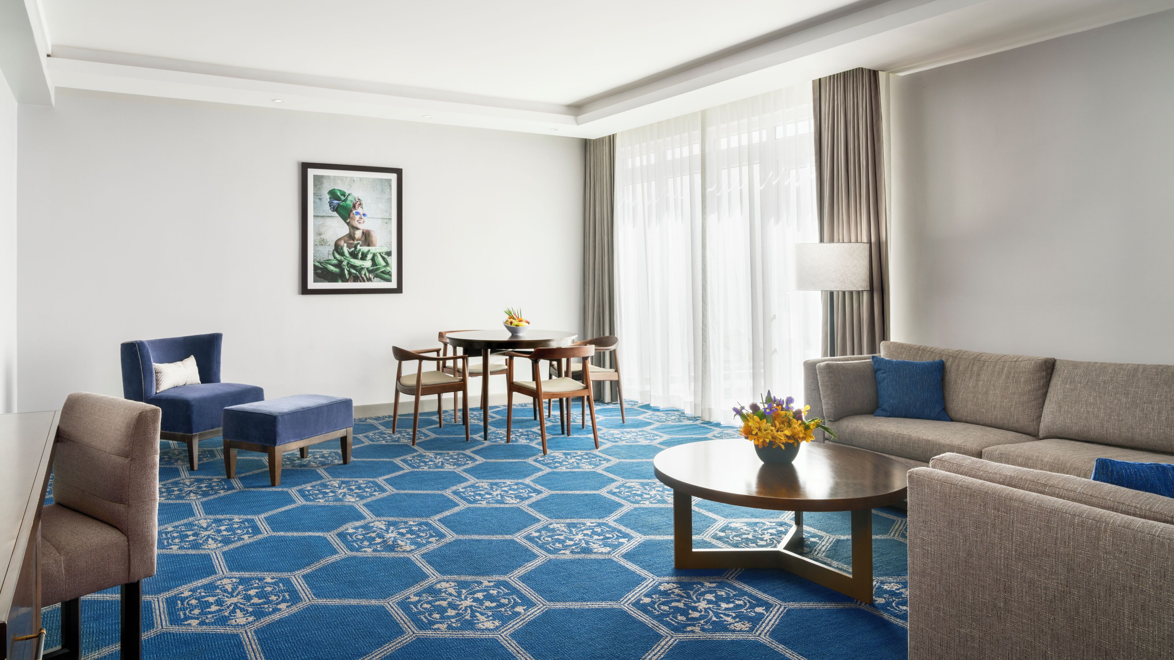Una habitación de hotel con alfombra azul hexagonal, un sofá, una silla, una mesa de comedor con sillas, un cuadro en la pared y una lámpara.