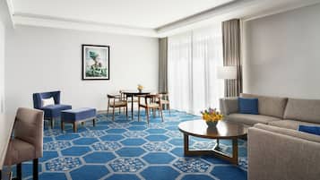 Una habitación de hotel con alfombra azul hexagonal, un sofá, una silla, una mesa de comedor con sillas, un cuadro en la pared y una lámpara.