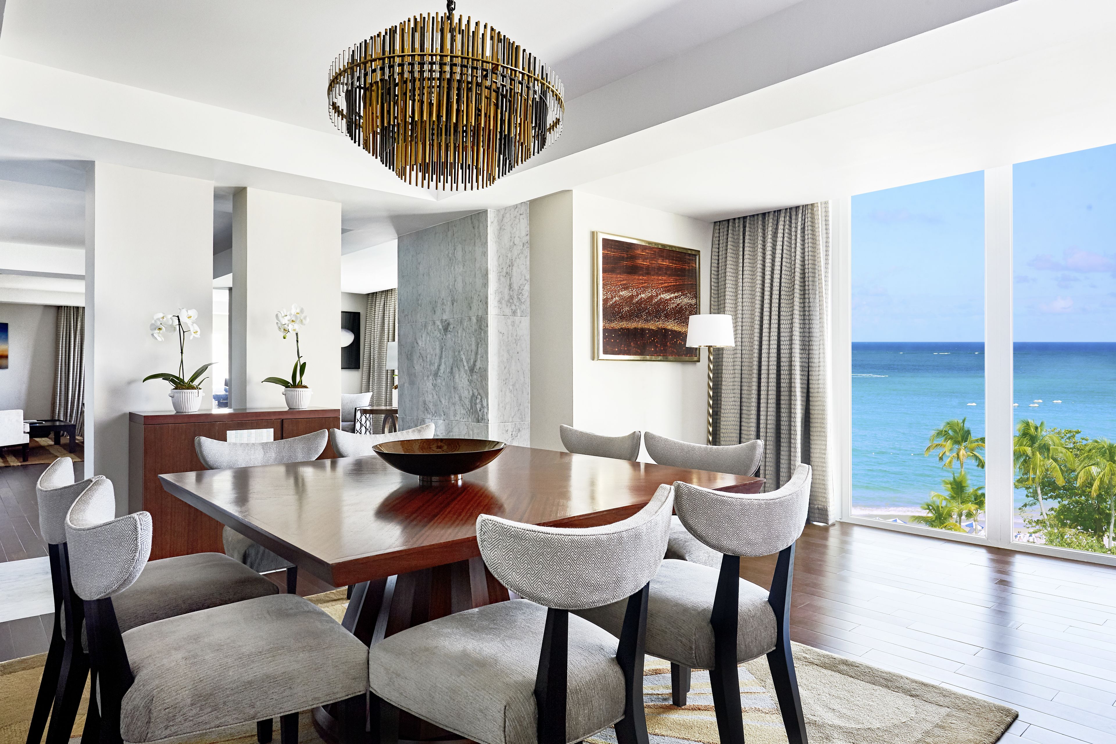 A modern dining area with a large wooden table, six upholstered chairs, a decorative bowl, and a view of the ocean through large windows.