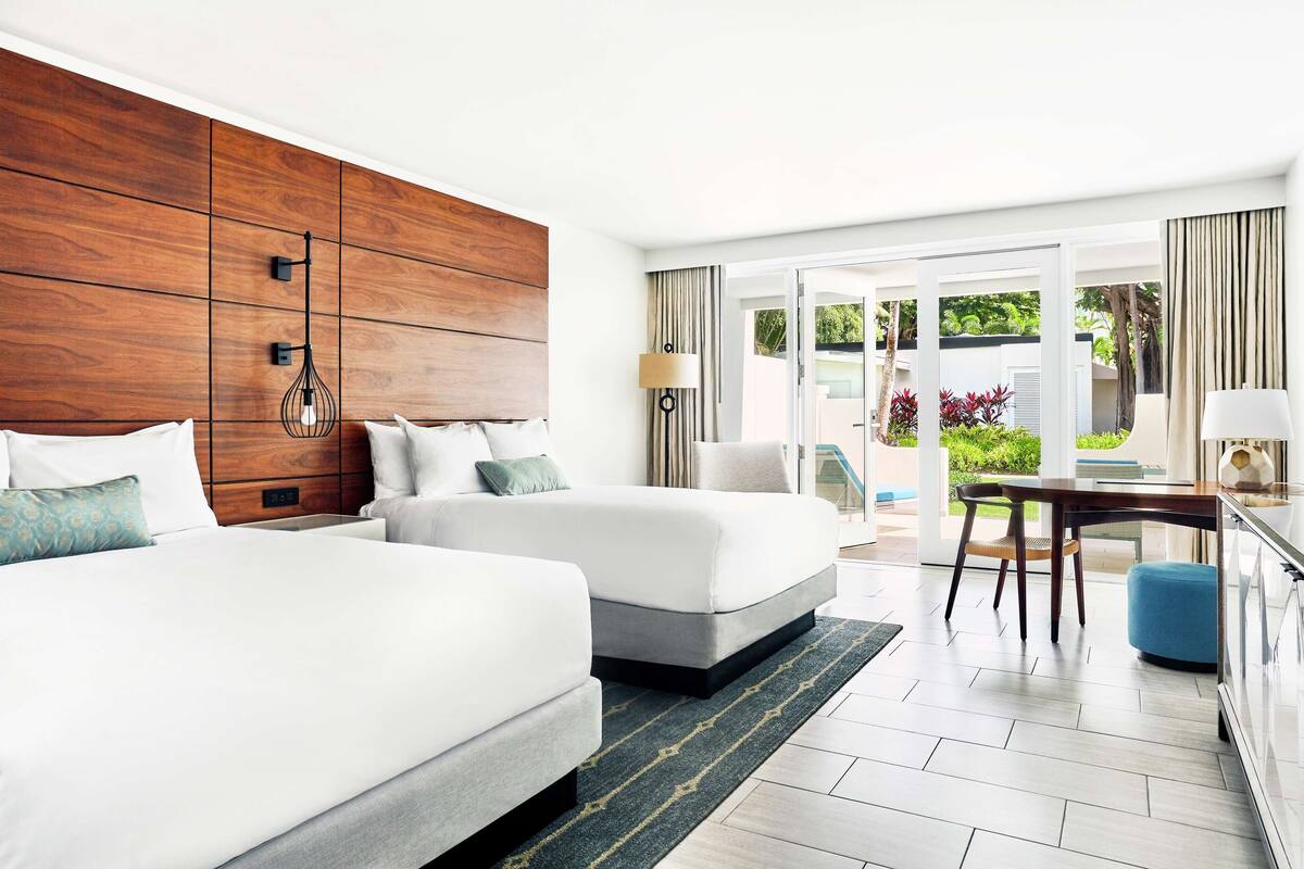 Une chambre d’hôtel avec deux lits, une tête de lit en bois, un coin repas avec une table et des chaises, et une vue sur l’extérieur.