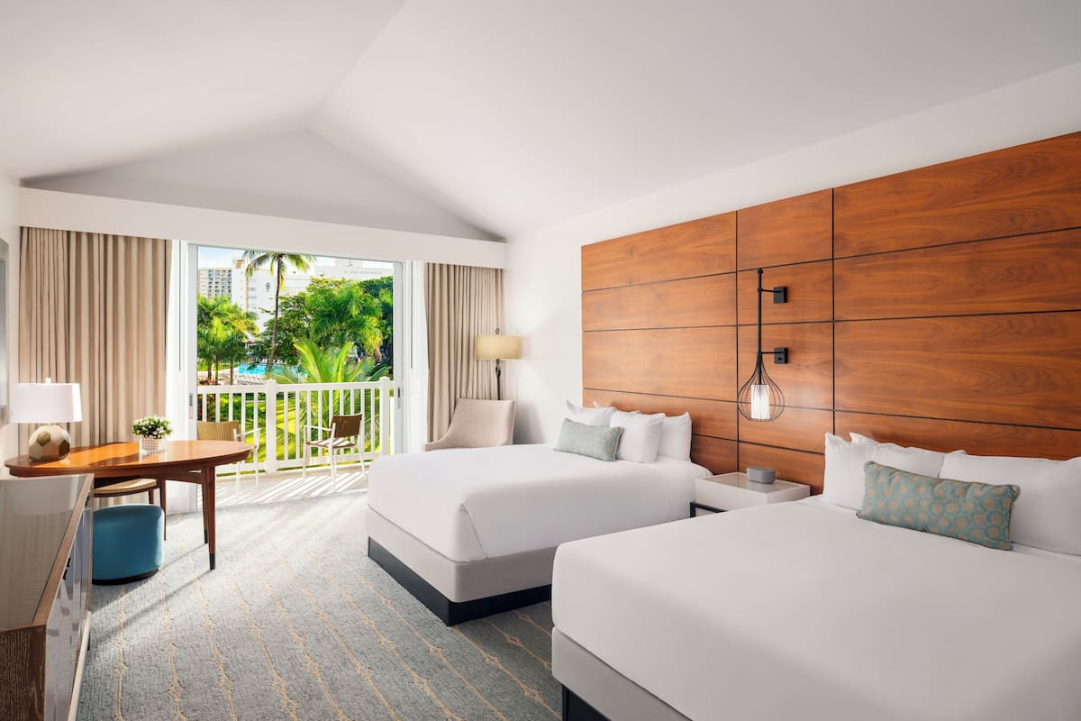 A hotel room with two beds, a desk, and a view of a pool and greenery.