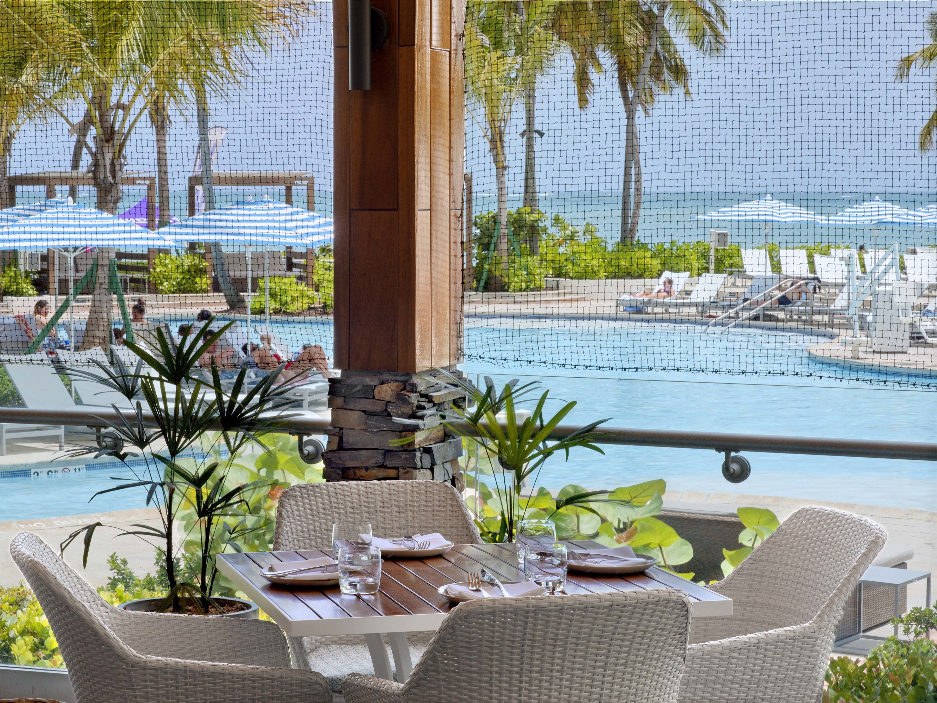 A poolside dining area with wicker furniture, a stone pillar, and a view of the pool and beach.
