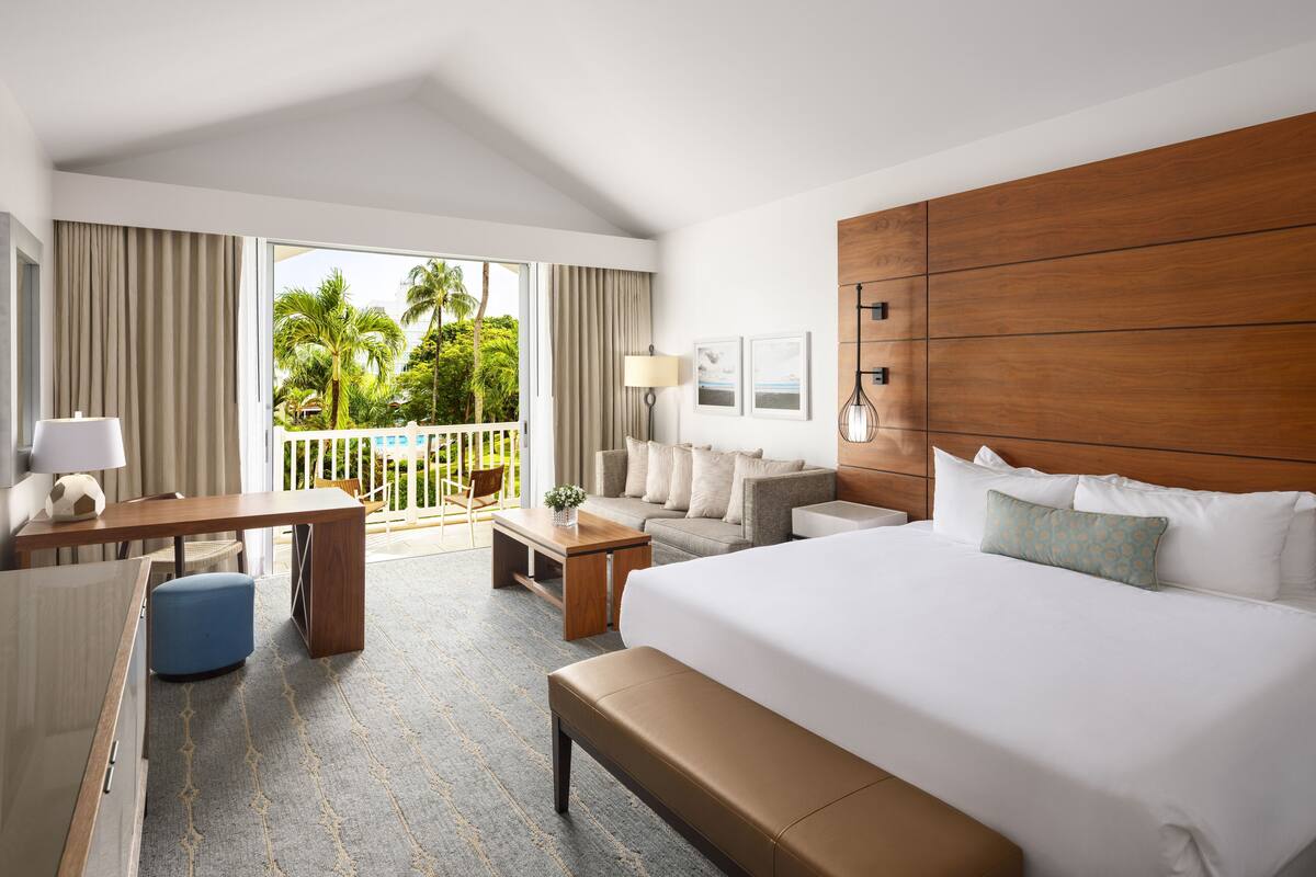 Une chambre d’hôtel avec un grand lit, un bureau, une chaise, un canapé et un balcon offrant une vue sur les palmiers.