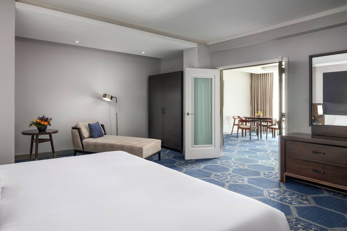 Une chambre d’hôtel avec un grand lit, un bureau avec une lampe, une chaise, une petite table avec des fleurs et une armoire.