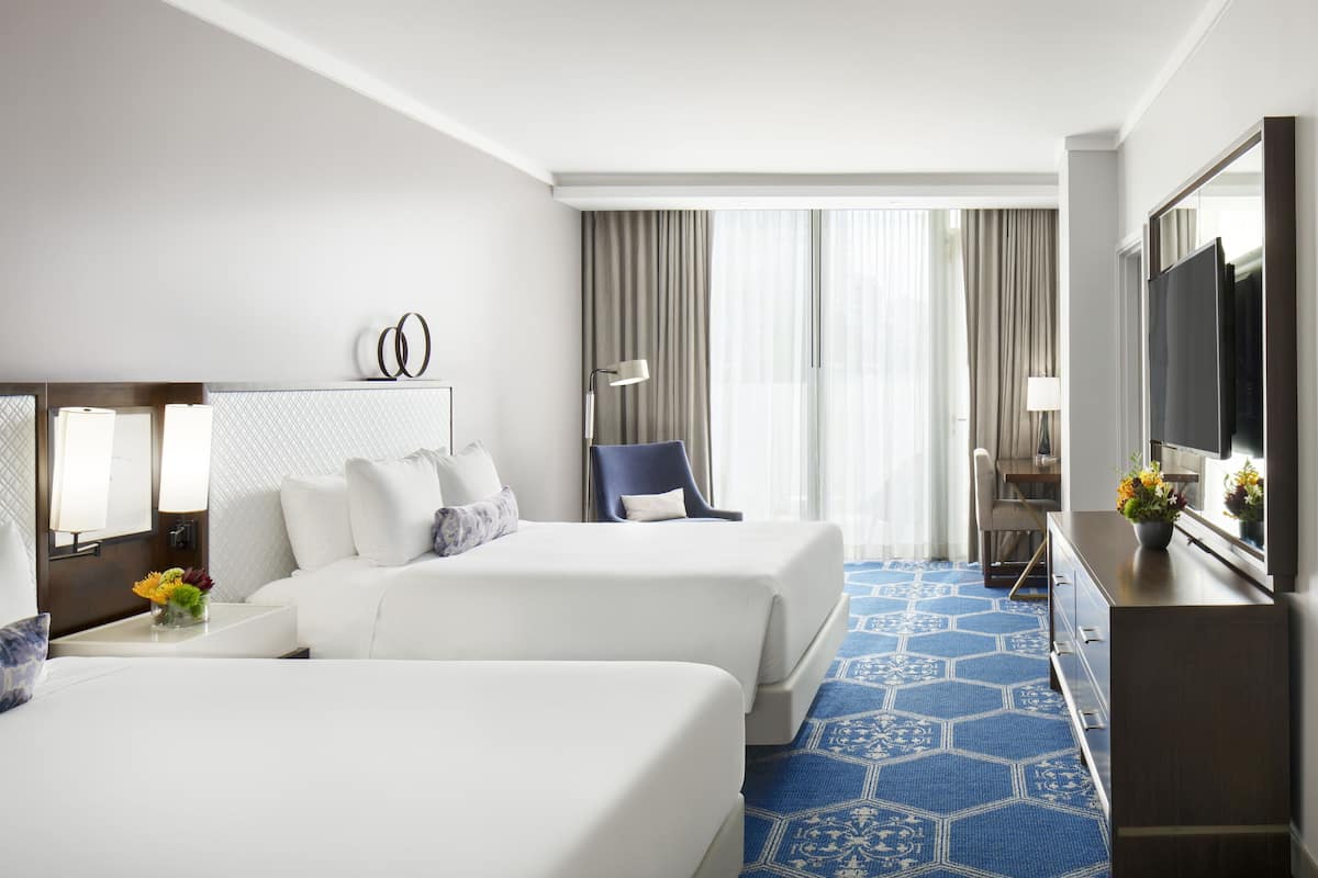 Une chambre d’hôtel avec deux lits, un bureau, une chaise, une télévision et une grande fenêtre avec des rideaux.