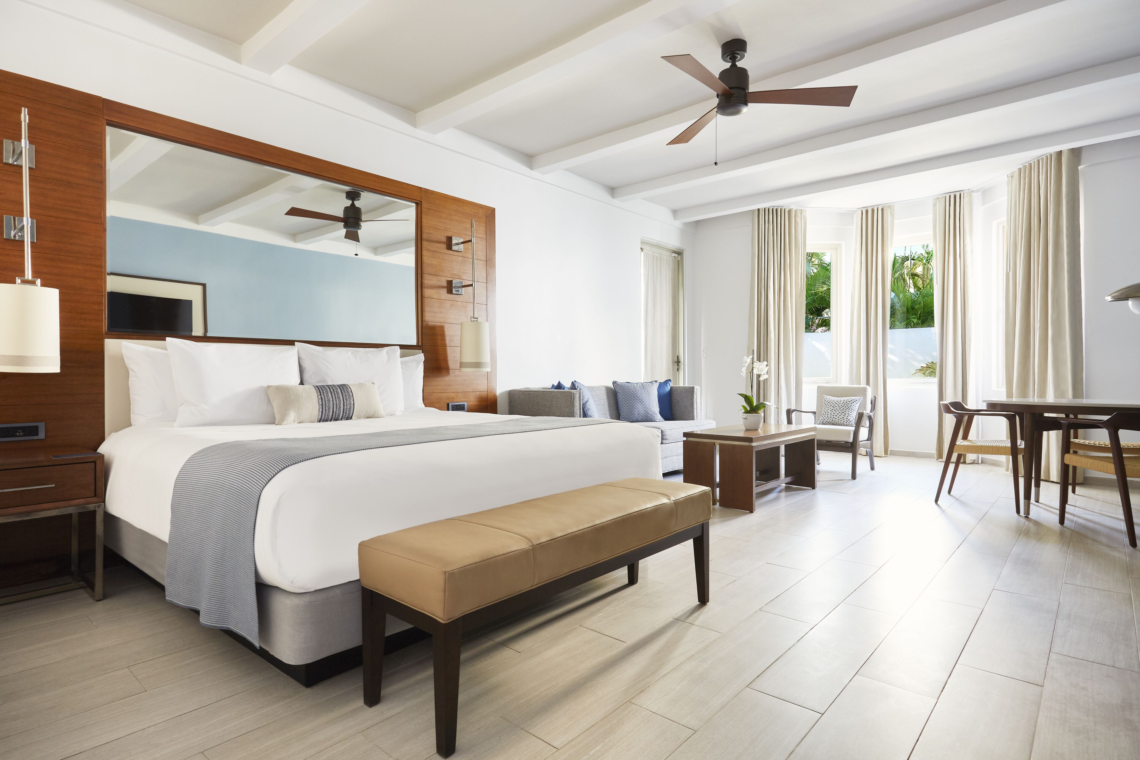 A modern hotel room with a large bed, a bench, a desk, and a balcony with curtains.
