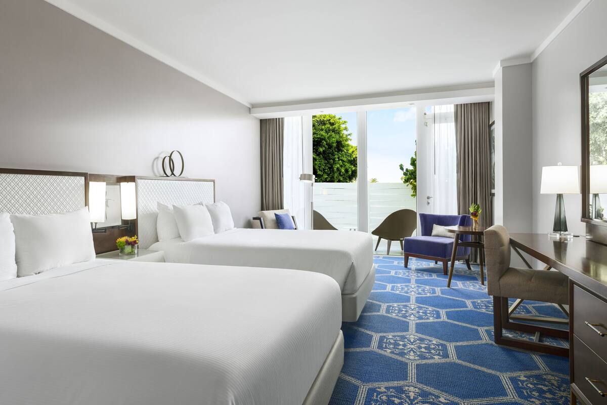 Une chambre d’hôtel avec deux grands lits, un bureau et une chaise. La fenêtre offre une vue sur la verdure.