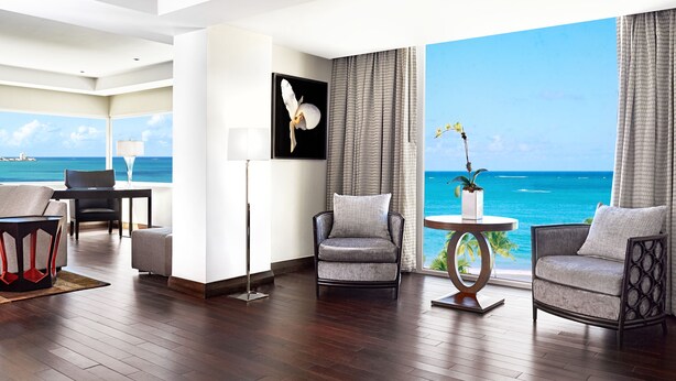A modern hotel room with a large window offering a view of the ocean, a seating area with two armchairs and a table, and a framed artwork on the wall. - Fairmont El San Juan Hotel (Carolina)