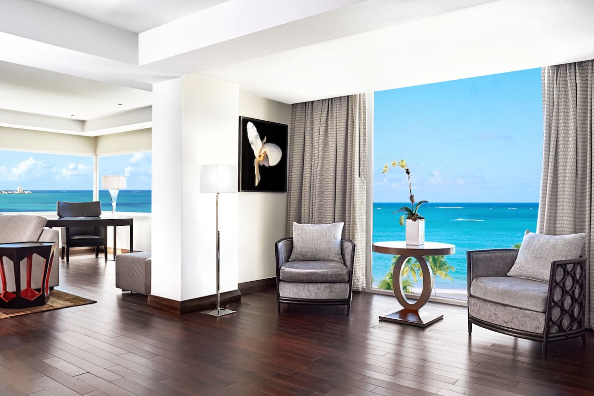 A modern hotel room with a large window offering a view of the ocean, a seating area with two armchairs and a table, and a framed artwork on the wall.