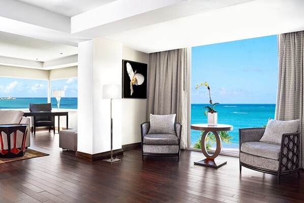 Une chambre d’hôtel moderne dotée d’une grande fenêtre donnant sur l’océan, d’un coin salon avec deux fauteuils et une table, ainsi que d’une œuvre d’art encadrée accrochée au mur.