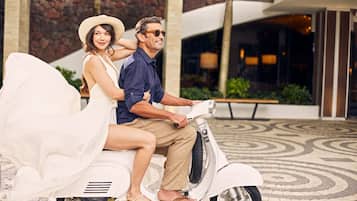Una pareja en un scooter blanco frente a un edificio con fachada de piedra y suelo estampado.