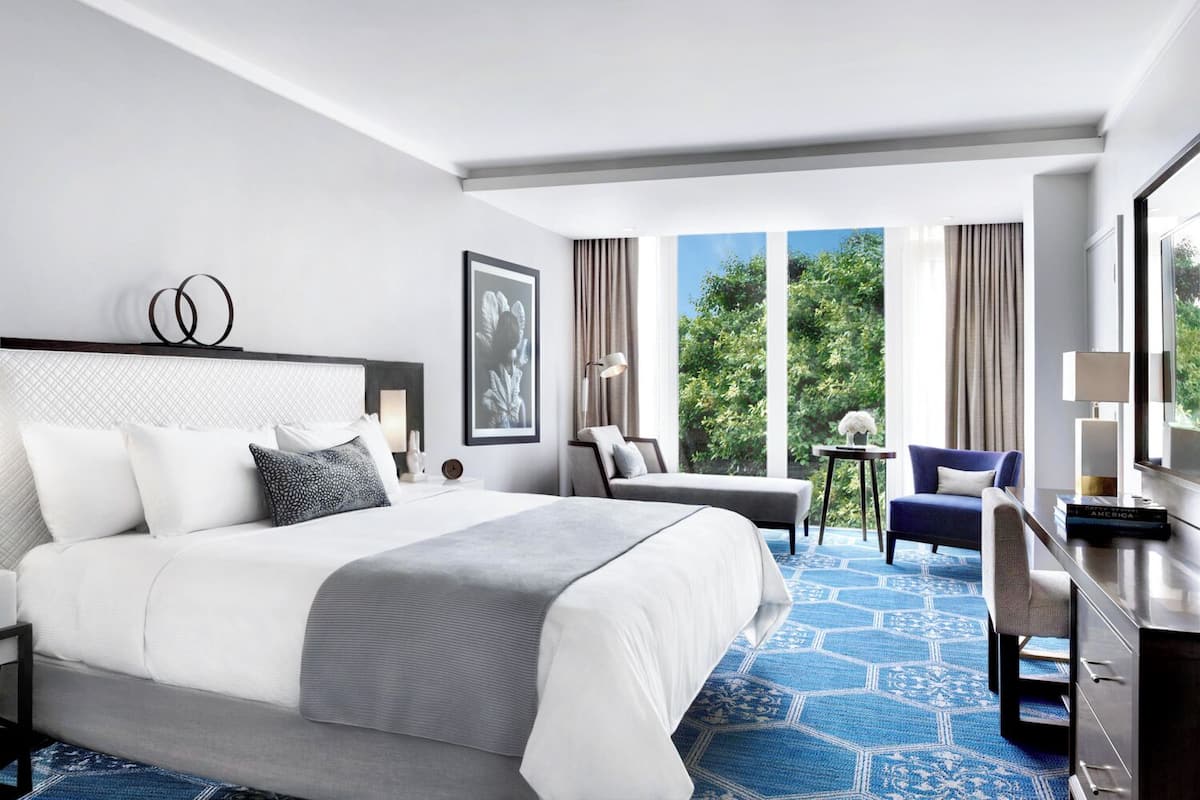 Une chambre d’hôtel moderne dotée d’un grand lit, d’un coin salon avec un canapé et des fauteuils, d’une petite table et offrant une vue sur la verdure grâce à de grandes fenêtres.