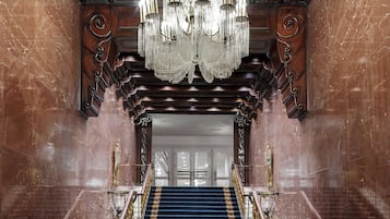 A grand staircase with a blue carpet and golden balusters, leading to a room with a large chandelier and ornate wood paneling.